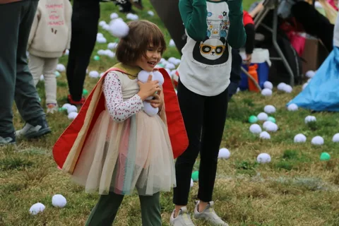 A child dressed as a princess with a red cape gathers white balls in her arms in the park.