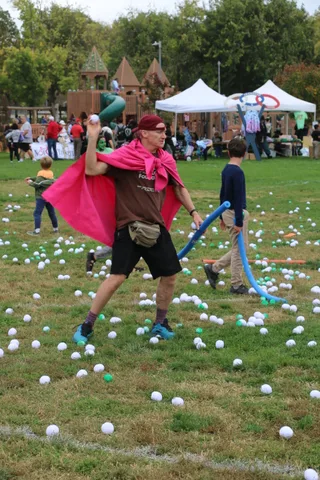 A man in a red cape raises a foam noodle over children on the grass.