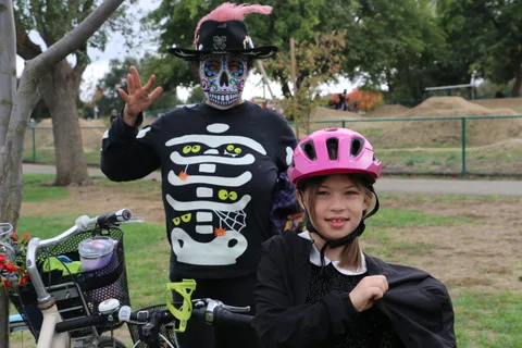 A man with a painted skull face waves beside a child in a pink helmet near parked bikes.