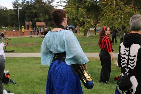 A performer in a teal cape carries a trombone while walking away.