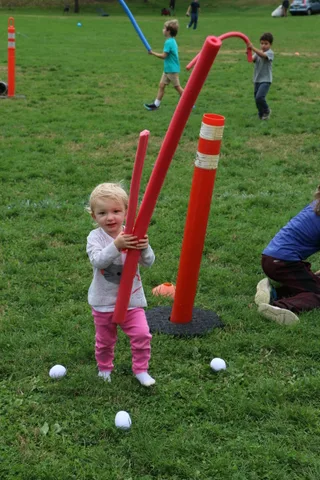 A toddler swings a striped foam pole near an orange safety cone on the grass.