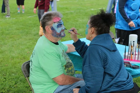 A volunteer paints a man's face blue and gray at a festival makeup booth.
