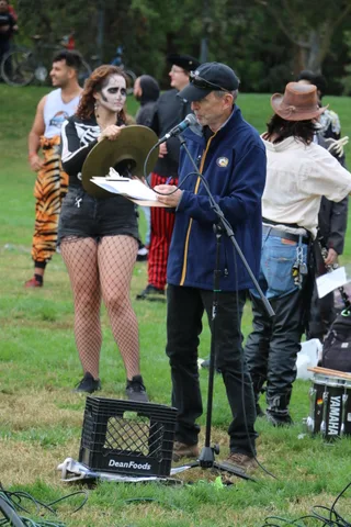 Costumed performers gather at the microphone, including a woman in skull makeup with cymbals.