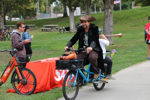 A cyclist dressed as a pirate rides a cargo bike along a park path.