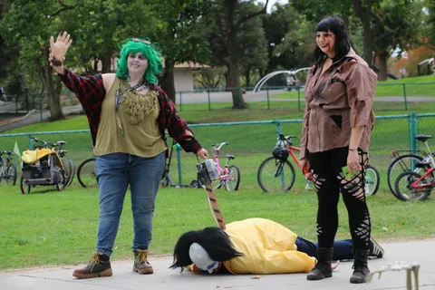 Two zombie performers stand over a costumed figure lying in the grass.