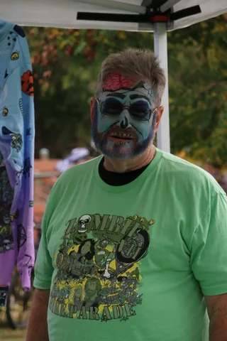 A man's face is painted in detailed grey zombie makeup with dark, hollow eyes.