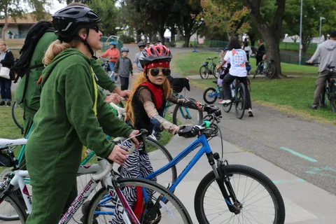 A man in a green costume walks his bike beside a woman in zombie makeup.