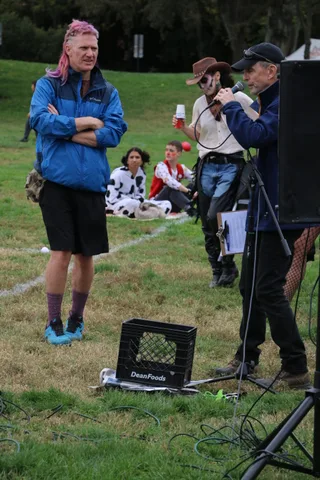 A man with arms crossed listens as another man speaks into a microphone.