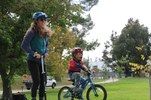 A woman rides a scooter while a boy pedals a bike behind her in the park.