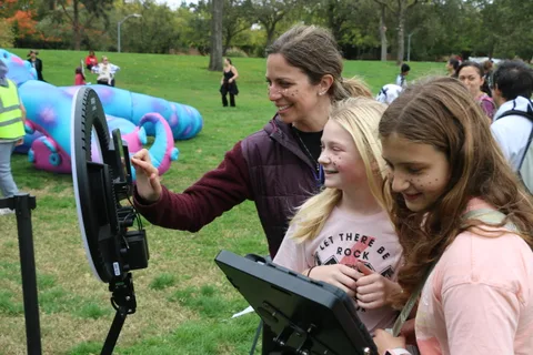 A woman waves with two laughing girls in front of the photo booth ring light.