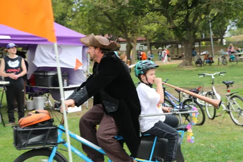 A costumed rider blows a horn while cycling past festival booths.