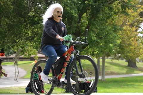 A woman in ghostly white face paint rides a decorated bicycle through the park.