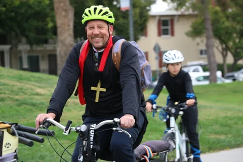A man in a neon helmet and priest-style costume rides with a child close behind.
