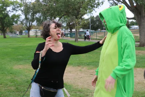 A woman in black with mouth open reaches toward a person in a green frog costume.