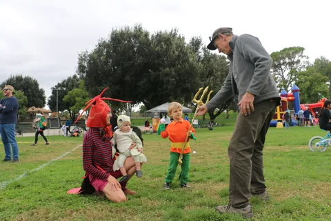 A man crouches down, pointing with a trident toy, near two children in costume.