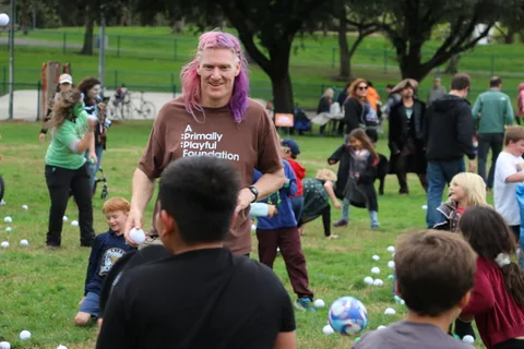 A man with pink hair wearing an A Primally Playful Foundation shirt looks up with an animated expression.