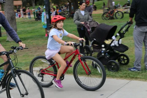 A girl rides a red bike as a family with a stroller looks on.