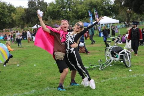 A man in a skeleton costume and pink cape celebrates with arms raised beside a laughing woman.