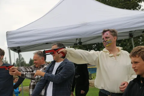 A man in cream face paint reacts as another aims a red toy blaster at him under a tent.