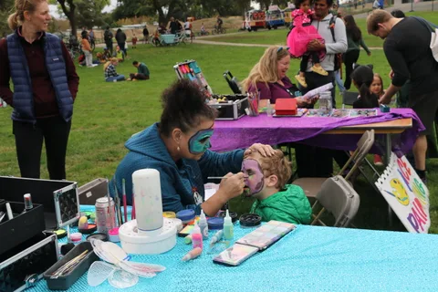 Two girls sit at a face painting table as one gets her design finished.
