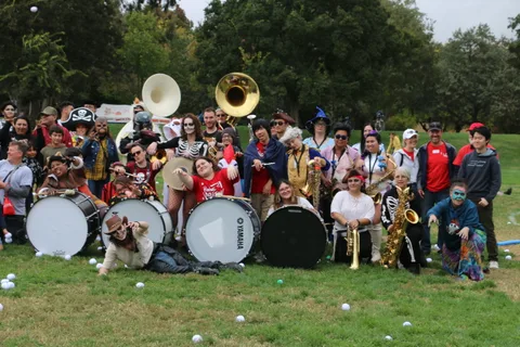 Marching band members with brass instruments and drums pose together outdoors.