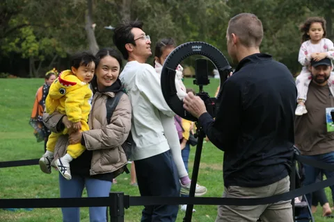 A mother holds her daughter, dressed as a chick, while a photographer captures the moment.