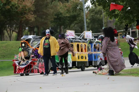 A Halloween-decorated parade train sits parked on a path as costumed staff walk nearby.