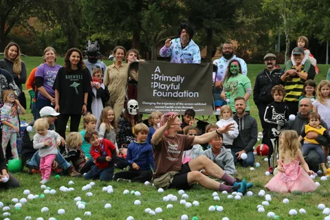 Costumed adults and children pose in a group photo beside an event banner in the park.