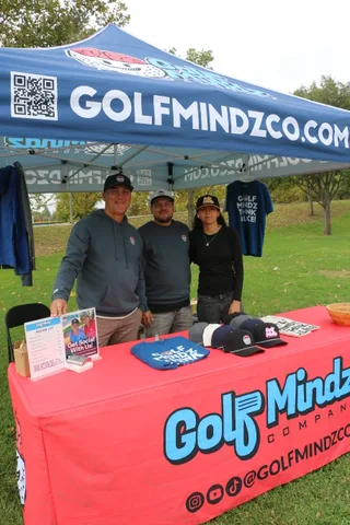 Three staff members pose under a GolfMindz sponsor tent with a QR code sign.