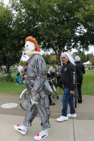 A clown-costumed figure walks in profile as a blurred person passes behind.