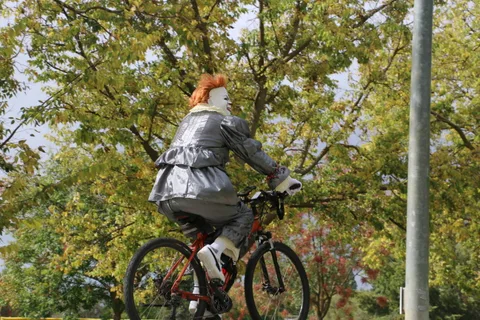A person in a silver costume rides a bicycle away from the camera under the trees.
