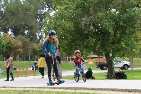 A mother on a scooter rides beside her son on a bike along a park path.