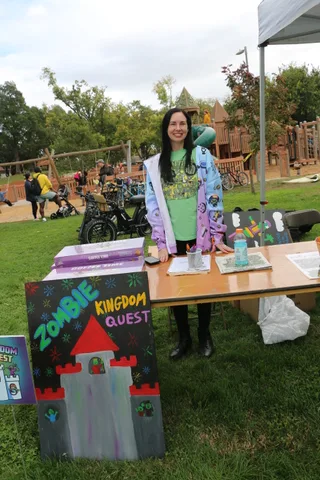 A woman in a green costume stands smiling in an open grassy area.