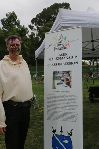 A man in zombie face paint stands beside a Davis Pentathlon laser marksmanship sign.