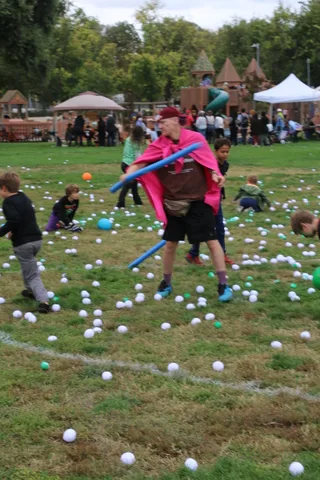 A man in a pink cape swings a foam noodle at children playing on the grass.