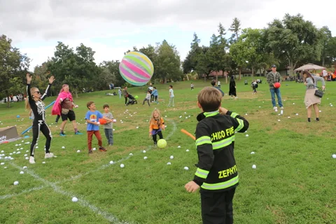 A woman in a flowing white costume reaches up to catch a ball above a park full of costumed kids.
