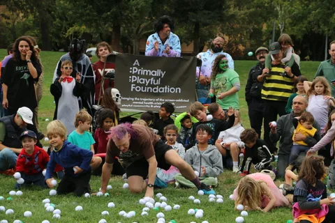 Children kneel and reach for scattered white balls in front of a crowd and event banner.