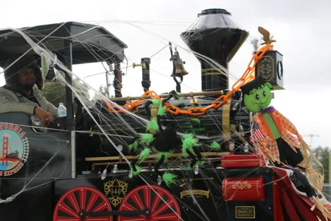 A decorated parade train with cobweb decor waits on the park path with its driver.