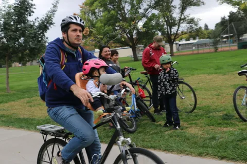 A father rides with a child on the front of his bike as onlookers watch.