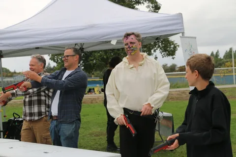 A man with skull face paint stands among a group aiming toy blasters under a tent.