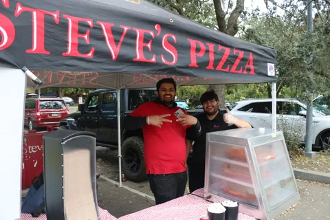 Two smiling staff give a thumbs up at the Steve's Pizza food booth.