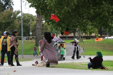 Costumed figures walk past a small decorated parade train on a park path.