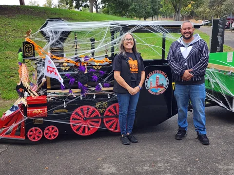 Two people pose beside a black trolley float decorated with spiderwebs and red wheels.