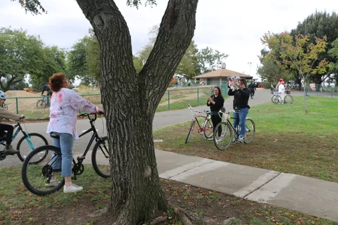 A cyclist walks a bike near a tree while another rider waves in the distance.