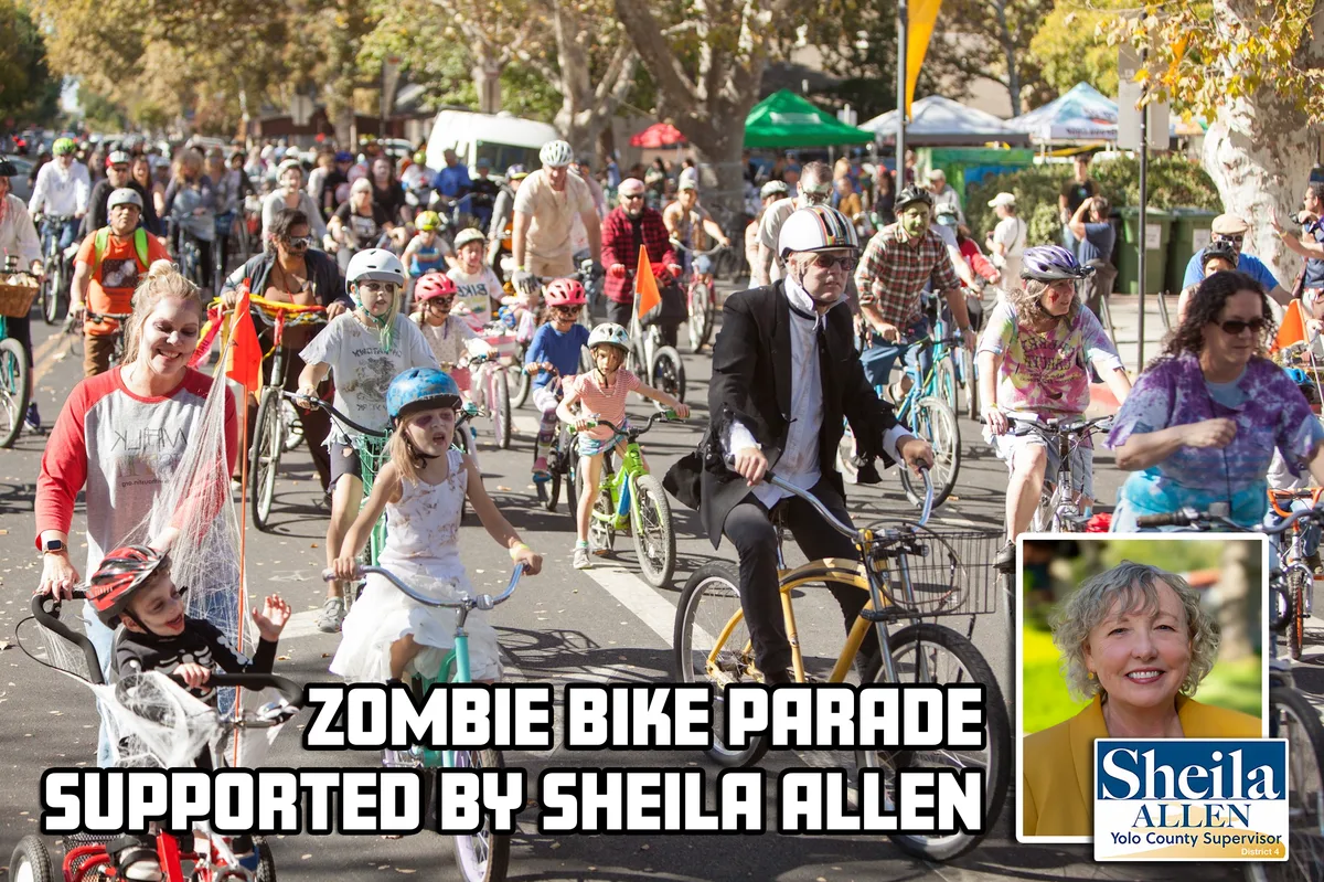 A crowd of costumed cyclists, including children in zombie makeup, ride together down a tree-lined street.