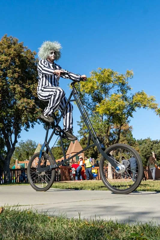 A tall novelty bicycle stands parked on the grass near the festival booths.