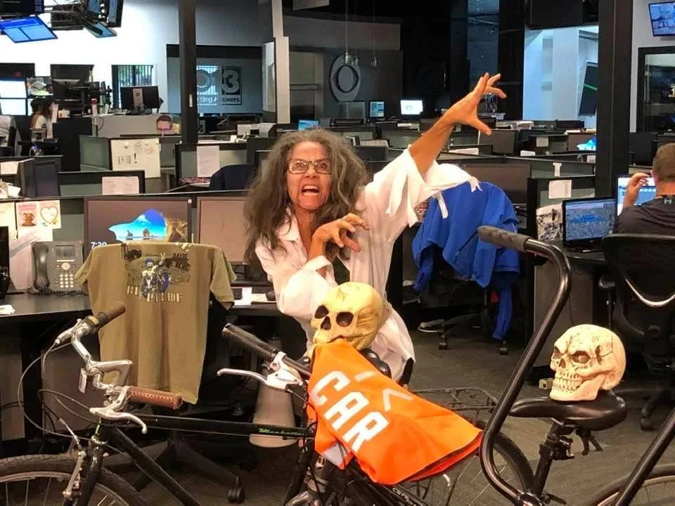 A woman in zombie makeup reaches toward the camera at an office desk.