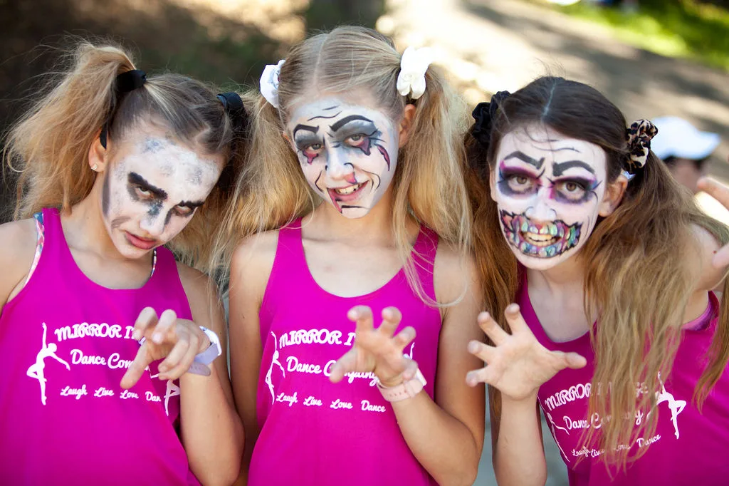 Three girls in zombie face paint and pink dance shirts pose together.