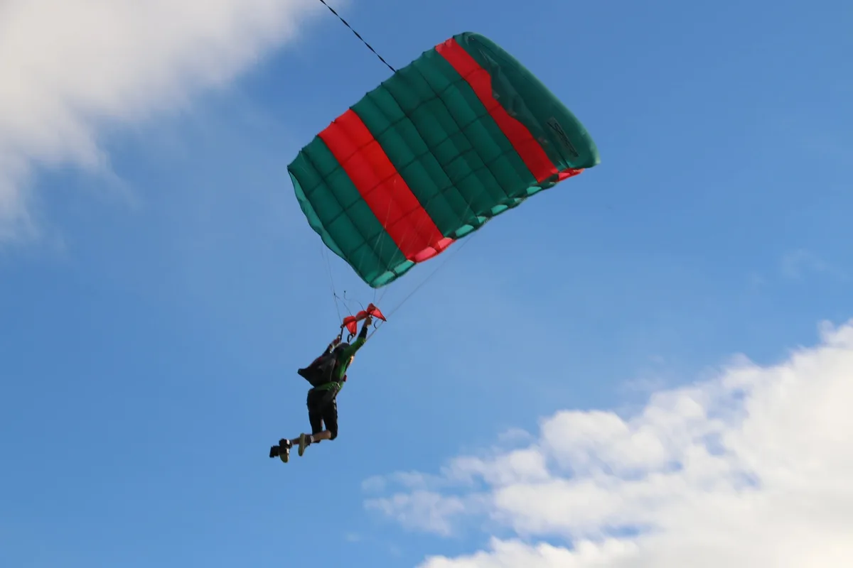 A skydiver descends under a green and red parachute against a clear blue sky.