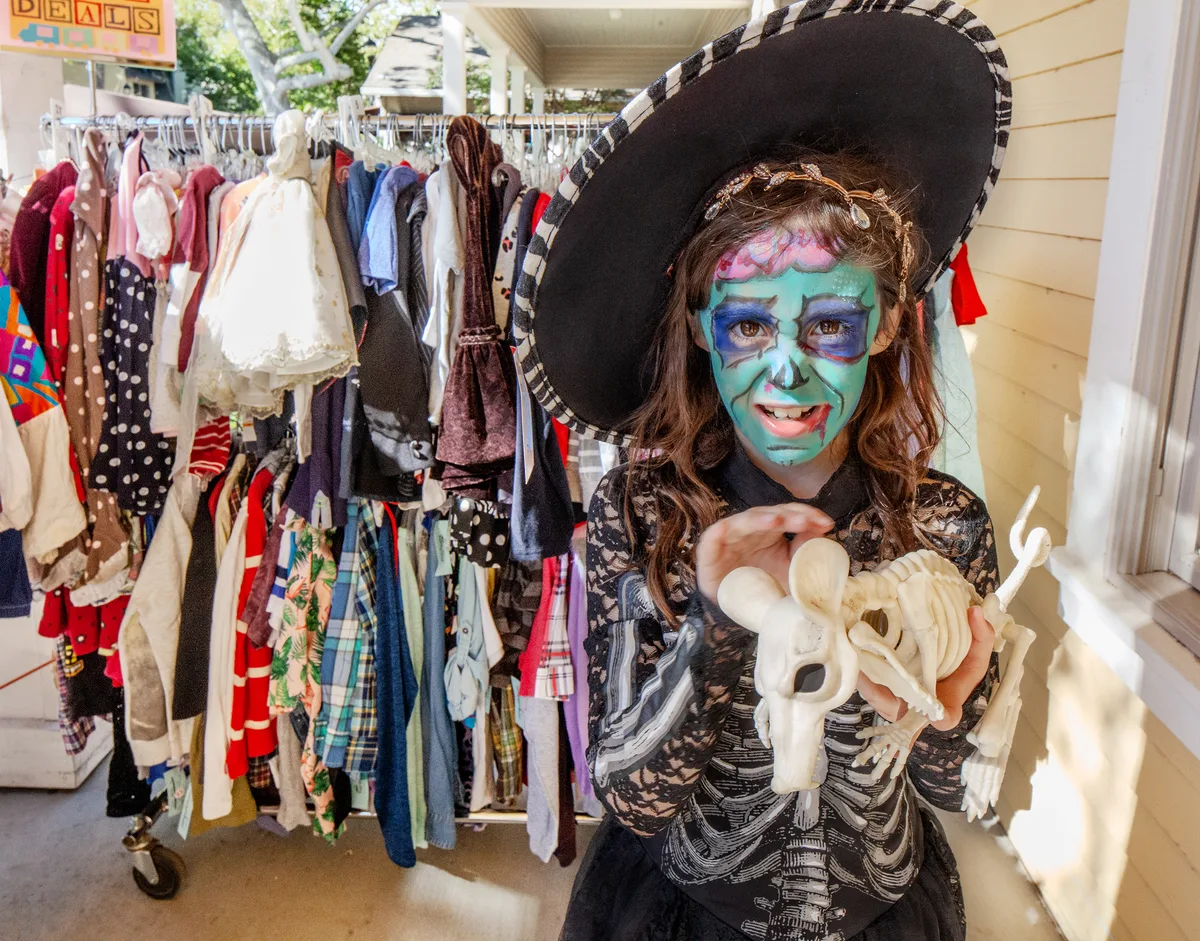A girl in a witch hat and skull face paint holds a toy skeleton at a clothing rack.